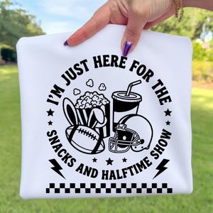 May include: White sweatshirt with a black graphic that reads "I'M JUST HERE FOR THE SNACKS AND HALFTIME SHOW." The graphic includes a football, popcorn, a drink, and a football helmet. The bottom of the sweatshirt has a black and white checkered pattern.