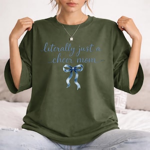 May include: Olive green t-shirt with the text "literally just a cheer mom" in blue script. A blue and white watercolor bow graphic is below the text. The shirt has short sleeves and a crew neck.