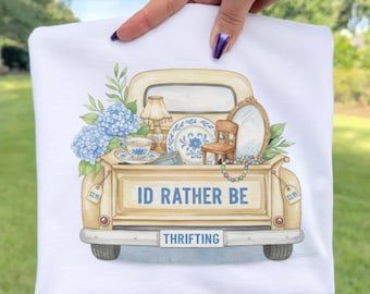 Könnte beinhalten: Weißes T-Shirt mit einer Aquarellillustration einer alten Pick-up-Ladefläche, die mit Antiquitäten gefüllt ist. Die Ladefläche trägt den Text "ID RATHER BE THRIFTING". Das Design umfasst blaue Hortensien, eine Teetasse und einen kleinen Stuhl.
