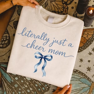 May include: Cream-colored sweatshirt with the text "literally just a cheer mom" in light blue script. A blue watercolor bow is below the text. The sweatshirt is held up by hands.