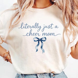 May include: Cream-colored t-shirt with the text "literally just a cheer mom" in blue script. A blue ribbon graphic is centered below the text. The shirt is made of a soft material.
