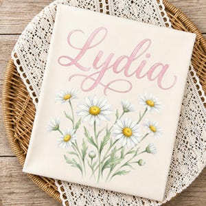May include: A canvas print featuring the name "Lydia" in pink script above a watercolor illustration of daisies. The daisies have white petals and yellow centers, with green stems and leaves. The print is on a beige background.