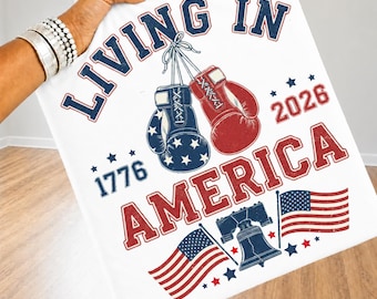 Könnte beinhalten: Ein weißer Stoffartikel mit einem Grafikdruck mit dem Text Living In America, 1776 und 2026. Das Design zeigt ein Paar zusammengebundene Boxhandschuhe, einen blauen mit weißen Sternen und einen roten, sowie zwei amerikanische Flaggen und eine Glocke.
