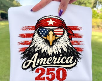 Könnte beinhalten: Weißes Sweatshirt mit einem patriotischen Adler-Design. Der Adler trägt eine Sonnenbrille mit dem Design der amerikanischen Flagge und ein rotes Bandana. Die Worte "America 250" sind unter dem Adler in einer stilisierten Schriftart aufgedruckt.