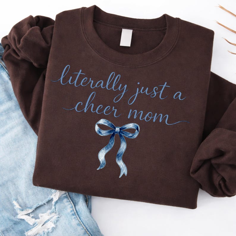 May include: A brown sweatshirt with the text "literally just a cheer mom" in blue script. A blue ribbon graphic is below the text. The sweatshirt is folded on top of blue jeans.