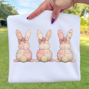 May include: White fabric featuring three patchwork bunny rabbits. Each bunny has a pink bow, patterned ears, and a round, textured tail. The bunnies are decorated with pink and cream-colored patterns.