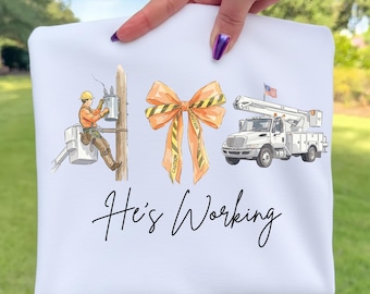 Könnte beinhalten: Weißes Sweatshirt mit einer Grafik eines Elektrikers bei der Arbeit, einer orangefarbenen Schleife mit Vorsichtband und einem weißen Nutzfahrzeug mit amerikanischer Flagge. Der Text "He's Working" ist kursiv geschrieben.
