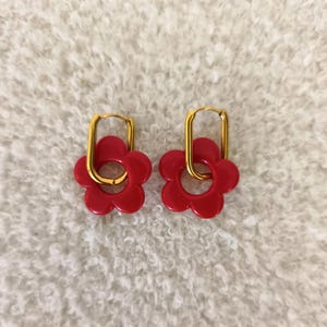 Peut inclure: Une paire de boucles d'oreilles rouges en forme de fleur avec des cerceaux dorés. Les boucles d'oreilles ont un design rétro en forme de marguerite. Les boucles d'oreilles sont présentées sur une surface blanche texturée.