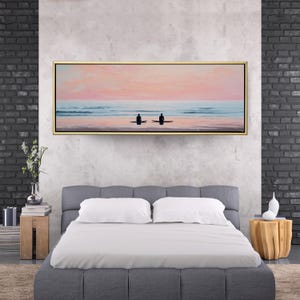 May include: A rectangular framed canvas art piece depicting two silhouetted figures on surfboards in the ocean at sunset. The sky is a blend of pink and peach hues, with the ocean in shades of blue. The frame is gold.