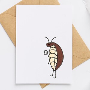 May include: A white greeting card with a cartoon cockroach illustration, holding a small cup. The card is on a brown envelope, with dried flowers and a white ribbon in the background.