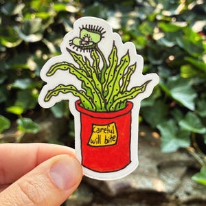 May include: A die-cut sticker featuring a green Venus flytrap plant with spotted leaves in a bright red pot. A yellow rectangular label on the front of the pot contains the handwritten black text "Careful will bite" in a casual, rounded font style.