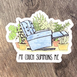 Puede incluir: Una pegatina con un sillón reclinable azul rodeado de plantas en macetas. El texto "MY COUCH SUMMONS ME" está escrito en negro. La pegatina tiene un borde blanco y está sobre una superficie de madera.