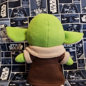 May include: A plush green character with large ears, wearing a brown robe with a tan collar. The character is set against a blue Star Wars-themed fabric background.