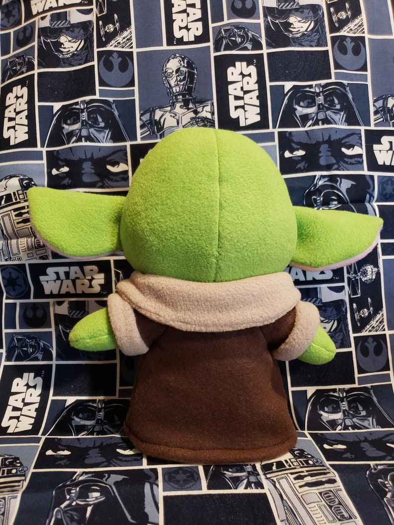 May include: A plush green character with large ears, wearing a brown robe with a tan collar. The character is set against a blue Star Wars-themed fabric background.