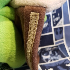 May include: Close-up of a plush toy's ear. The ear is brown with a beige trim and a beige fabric strip. The plush toy has a green body and a white inner ear. The background is a blue fabric with a Star Wars comic book design.