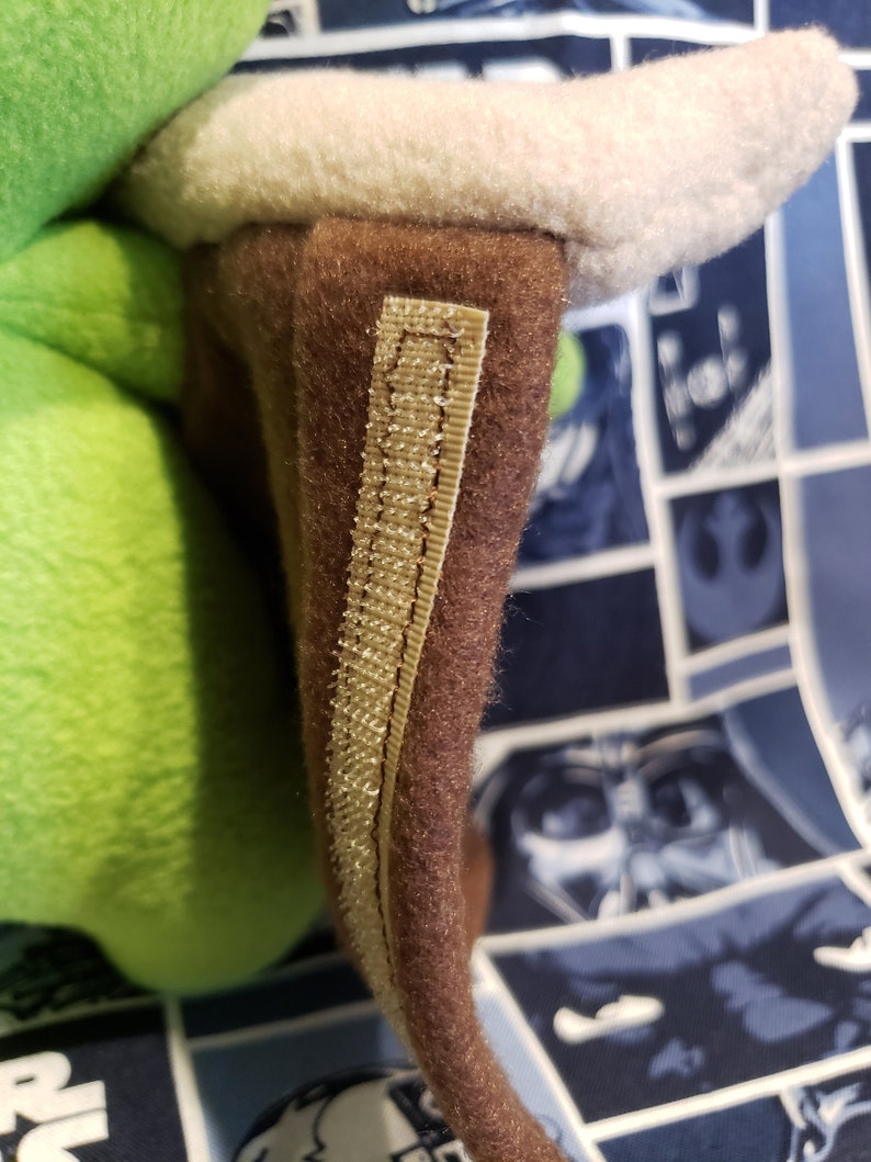 May include: Close-up of a plush toy's ear. The ear is brown with a beige trim and a beige fabric strip. The plush toy has a green body and a white inner ear. The background is a blue fabric with a Star Wars comic book design.