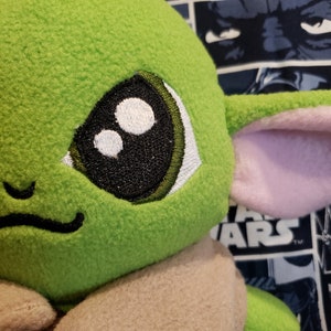 May include: Close-up of a plush green character with large black and white eyes, a small smile, and large ears with pink inner details. The character's body is a light tan color. The background features a Star Wars-themed fabric.