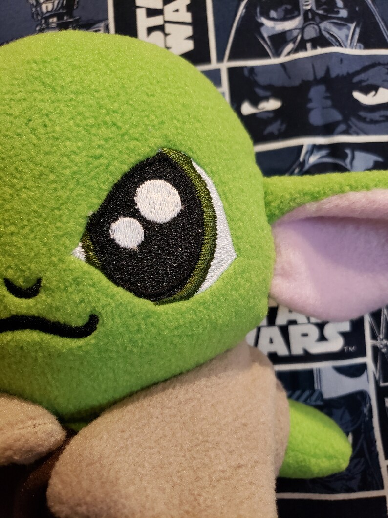 May include: Close-up of a plush green character with large black and white eyes, a small smile, and large ears with pink inner details. The character's body is a light tan color. The background features a Star Wars-themed fabric.