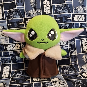 May include: A plush green character with large black eyes, pink-lined ears, and a brown coat. The character is set against a blue and white Star Wars-themed fabric background featuring various characters and the Star Wars logo.