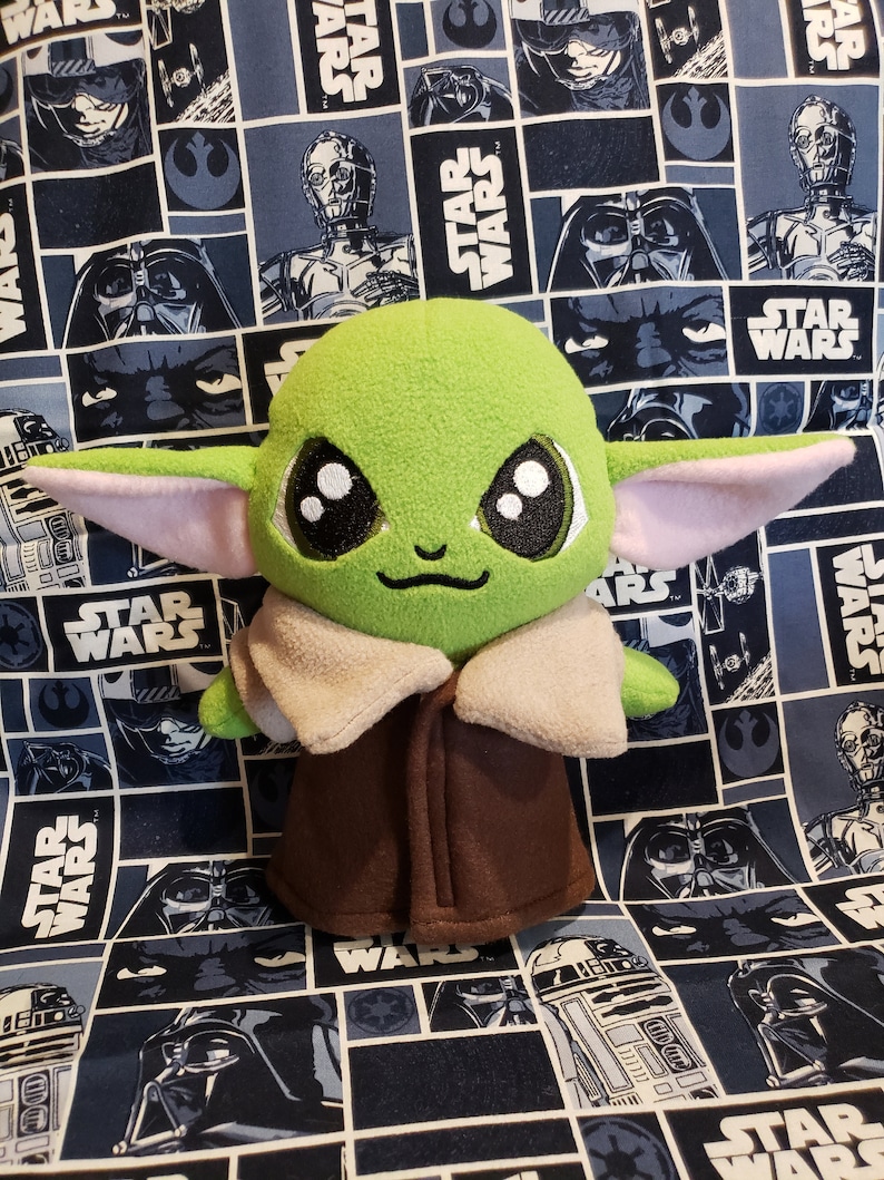 May include: A plush green character with large black eyes, pink-lined ears, and a brown coat. The character is set against a blue and white Star Wars-themed fabric background featuring various characters and the Star Wars logo.