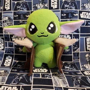 May include: A plush green Grogu doll with large black eyes, light pink ears, and a brown cloak. The doll is set against a blue Star Wars-themed fabric background featuring various characters and the Star Wars logo.
