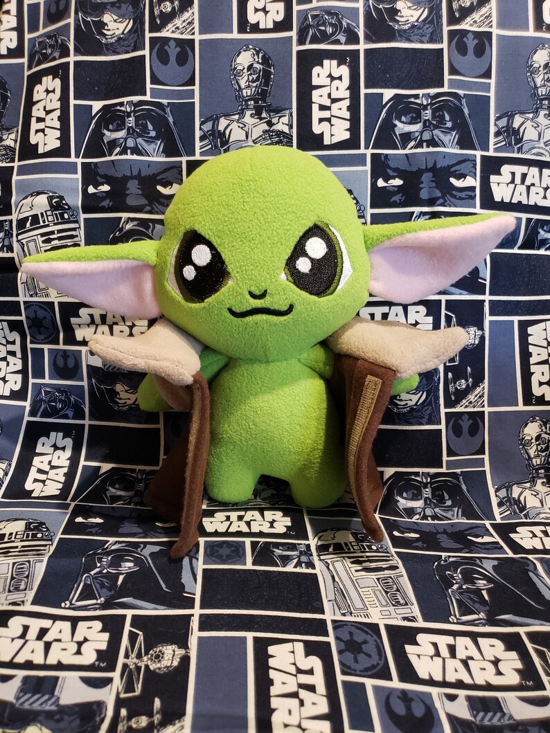 May include: A plush green Grogu doll with large black eyes, light pink ears, and a brown cloak. The doll is set against a blue Star Wars-themed fabric background featuring various characters and the Star Wars logo.