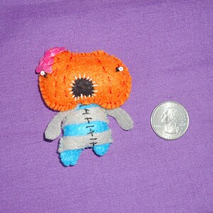 May include: A handmade felt doll with an orange head, a gray and blue striped body, and small arms and legs. The doll has a black mouth with white stitching and pink sequin accents. A U.S. quarter is next to the doll for size comparison.