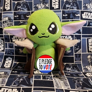 May include: A plush green character with large eyes and pointed ears, wearing a brown cloak. The character is set against a blue Star Wars-themed background. A circular button with the text "I PLEDGE TO VOTE" is in the foreground.
