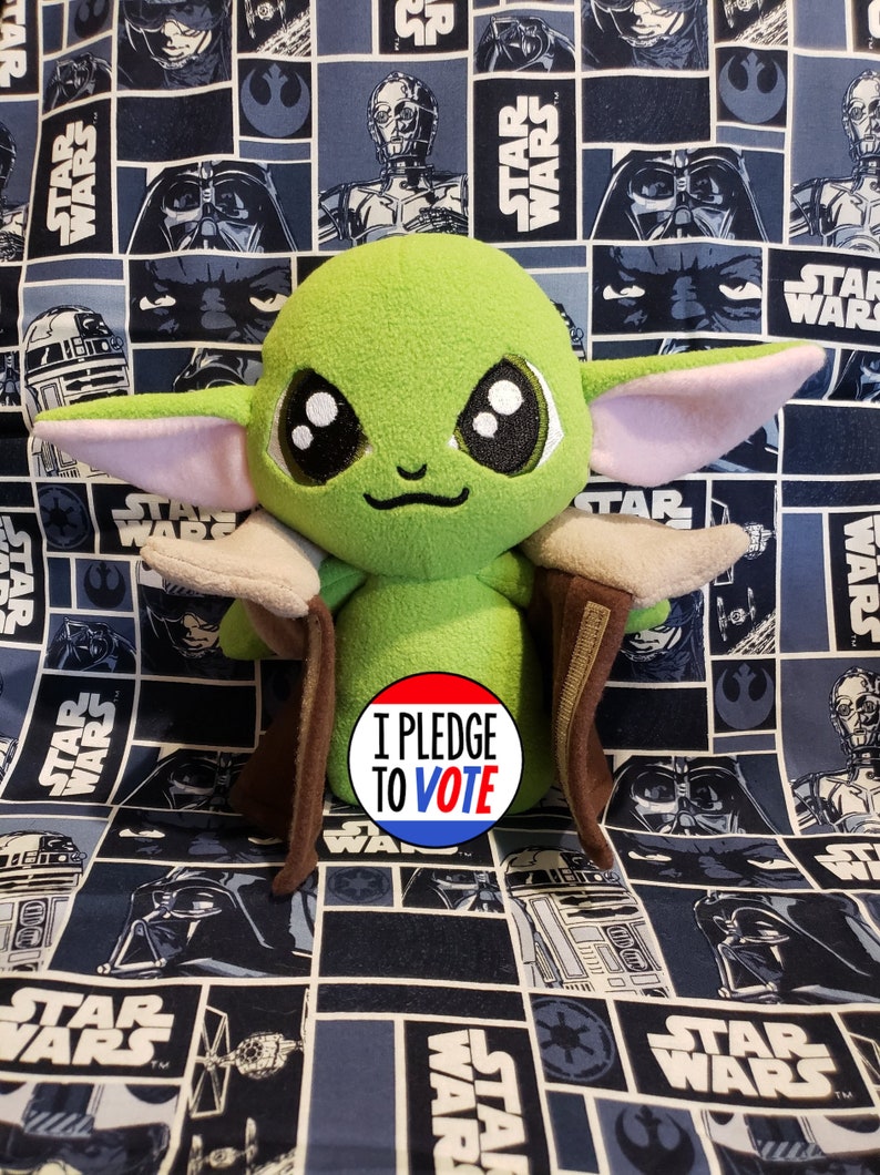 May include: A plush green character with large eyes and pointed ears, wearing a brown cloak. The character is set against a blue Star Wars-themed background. A circular button with the text "I PLEDGE TO VOTE" is in the foreground.