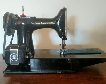 May include: A black Singer sewing machine with a metal foot pedal and a black and silver needle. The machine has a black belt and a black and silver motor. The machine is sitting on a wooden table.