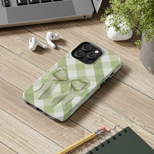May include: A phone case with a green and white gingham pattern and a watercolor bow design. The case is on a wooden surface with a laptop, earbuds, a pencil, and a notebook.