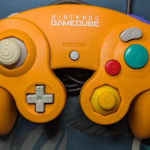 Peut inclure: Manette Nintendo GameCube orange avec des boutons blancs, gris, rouges, jaunes et turquoise. Le contrôleur porte les inscriptions "NINTENDO GAMECUBE" et "START/PAUSE". Accessoire de jeu classique.