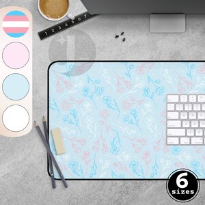 Peut inclure: Tapis de bureau bleu clair avec un motif floral rose, blanc et bleu. Le tapis a une bordure noire et un clavier blanc est partiellement visible. L'image montre également un crayon, une gomme et une tasse de café.