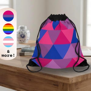 May include: A drawstring bag featuring a geometric pattern of triangles in shades of pink, purple, and blue. The bag has black cord straps and a gathered top closure. To the left, a vertical panel displays three circular pride flag icons and the text & more!
