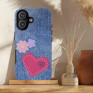 Puede incluir: Una funda de teléfono azul estilo denim con un corazón rosa y dos flores. El corazón está cosido con hilo blanco y tiene un pequeño detalle de botón. La funda tiene un recorte para la cámara y se muestra sobre un soporte de madera.