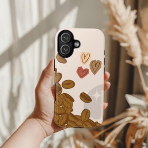 May include: A phone case with a beige background featuring a coffee theme. The design includes scattered coffee beans and three heart shapes in red, brown, and beige. The case is held in a hand.