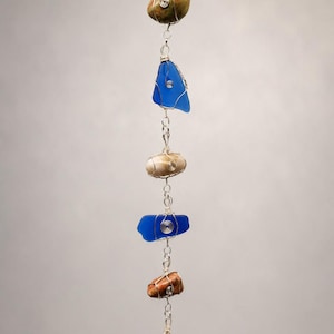 May include: A vertical hanging ornament featuring alternating pieces of smooth sea glass in vibrant blue and various natural stones in shades of brown, tan, and green, all individually wrapped in silver-toned wire and connected by metal links.