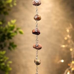 May include: A vertical hanging ornament featuring seven polished, multi-colored stones wrapped in silver-toned wire. Each stone is connected by silver-toned metal links, creating a linear chain structure that hangs from a single silver-toned ring.