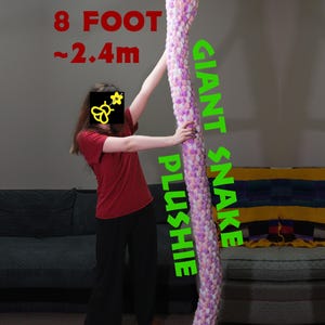 May include: A large, knitted snake plushie, measuring approximately 2.4 metres in length, in a mix of pink, purple, and white. The words "GIANT SNAKE PLUSHIE" are printed vertically. The text "8 FOOT ~2.4m" is printed in red.