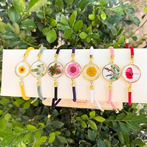 May include: A collection of seven gold-toned bracelets, each featuring a clear, circular pendant with a different pressed flower. The bracelets have colorful cords in yellow, green, black, purple, white, pink, and red.