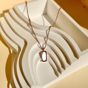 May include: A rose gold necklace with a rectangular pendant featuring a white, iridescent stone. The necklace is displayed on a white, layered, abstract tray, with shadows adding depth. The chain is delicate and the pendant is centered.
