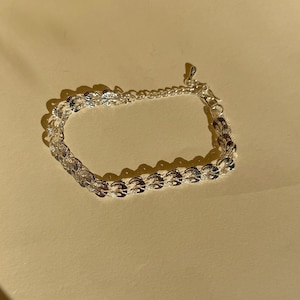 May include: A silver bracelet with a delicate chain design. The bracelet features a series of interconnected, textured links that catch the light. The adjustable chain allows for a customized fit. The jewelry is displayed on a neutral background.
