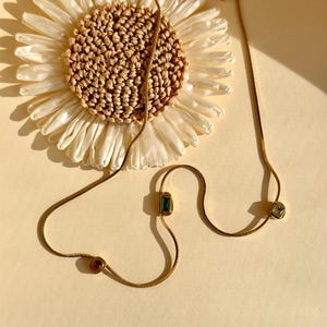 May include: Gold-toned necklaces with gemstone accents. One necklace features a rectangular emerald-green stone and a round, reddish-brown stone. Another necklace has a square, clear stone. The necklaces are displayed on a light yellow surface with a decorative flower.