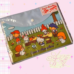 May include: A clear plastic document folder featuring a colorful Sanrio characters picnic illustration. The design shows Hello Kitty and friends on a green lawn with a white fence, a wooden table, and the text Sanrio characters printed in purple.