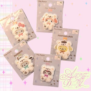 Peut inclure: Cinq écussons en relief Sanrio sur des cartes de support grises individuelles. Chaque écusson représente un personnage portant un costume d'ours blanc. Les personnages incluent Hello Kitty avec un nœud rose, My Melody, Pompompurin, Pochacco et Kuromi.