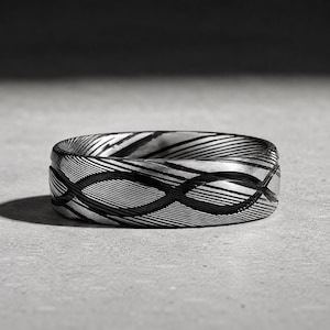 May include: A ring featuring a dark gray and black Damascus steel pattern with a distinct wavy, layered aesthetic. The metal surface displays a continuous, flowing design of alternating light and dark lines that wrap around the entire band.