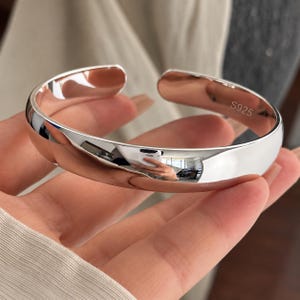 May include: A polished silver cuff bracelet with an open design. The interior surface of the metal band features the engraved text S925, indicating the sterling silver composition. The bracelet has a smooth, reflective, and curved exterior.