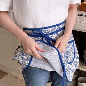 Puede incluir: Un delantal con estampado floral azul y blanco con ribete azul e interior a rayas. El delantal tiene cierre de lazo y dos bolsillos. El delantal lo sostiene una persona que lleva una camisa blanca y vaqueros azules.