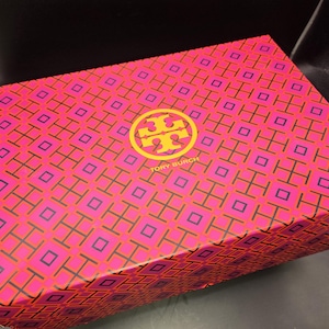 May include: A pink and magenta patterned box with a yellow logo and the text "TORY BURCH". The box has a repeating geometric design and is likely for a fashion item.