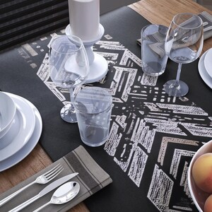 May include: A dining table set with white plates, bowls, and glasses. Silver cutlery rests on a grey napkin. A black table runner with a white geometric pattern is in the center. Peaches and grapes are in a bowl.